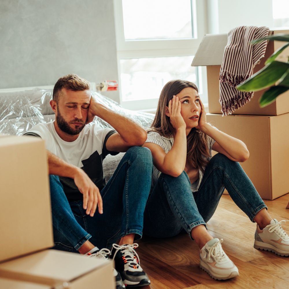 Frustrated couple sitting among moving boxes indoors