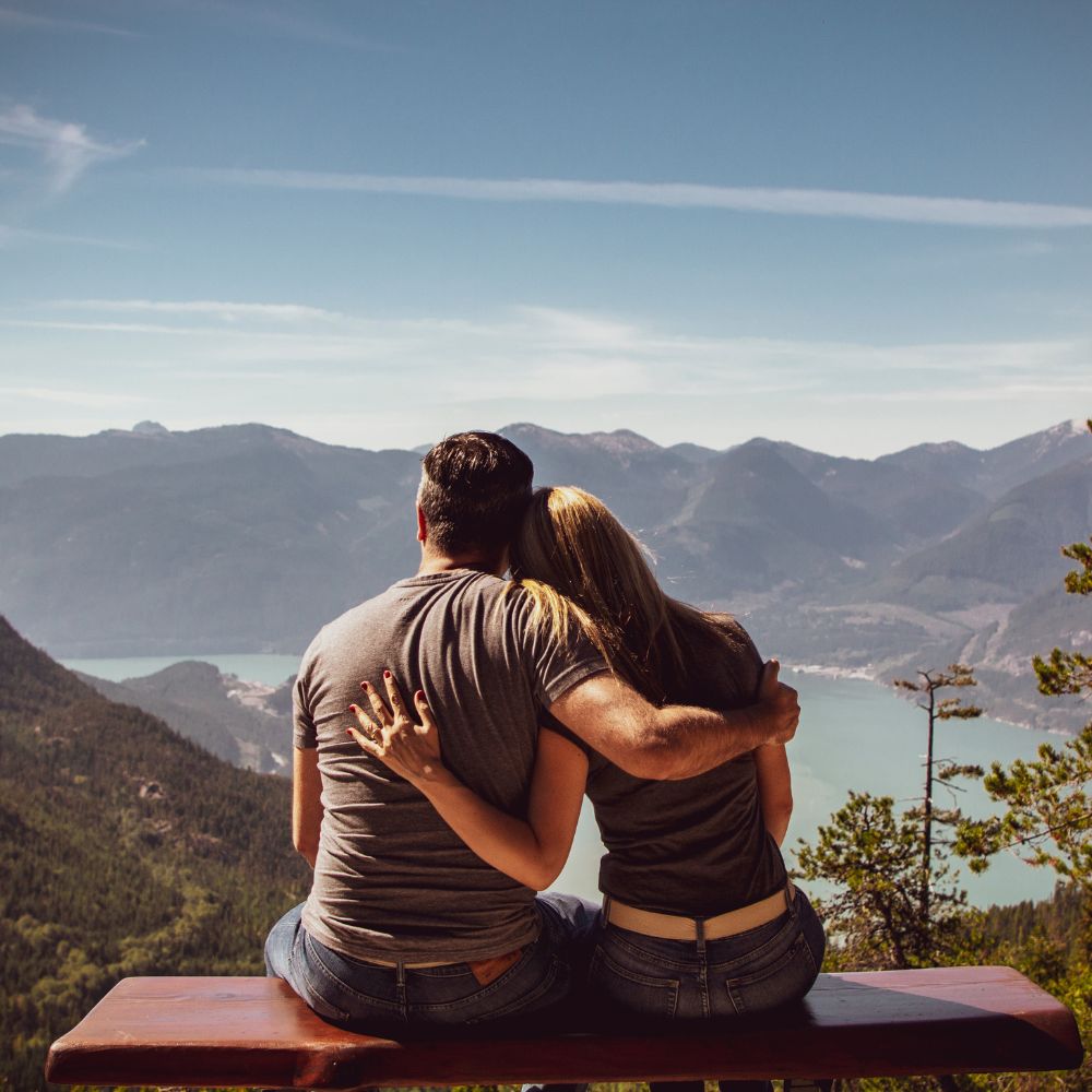 Couple sitting on bench hugging while admiring mountain lake view