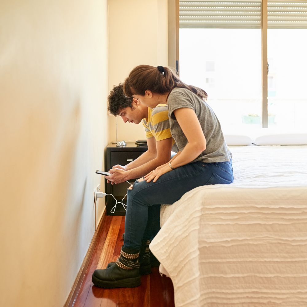 Young couple sitting on bed focused on phone charging near wall