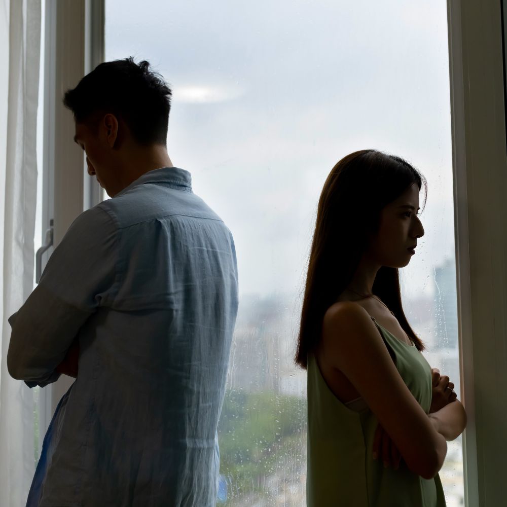 Couple standing back-to-back looking upset near rainy window
