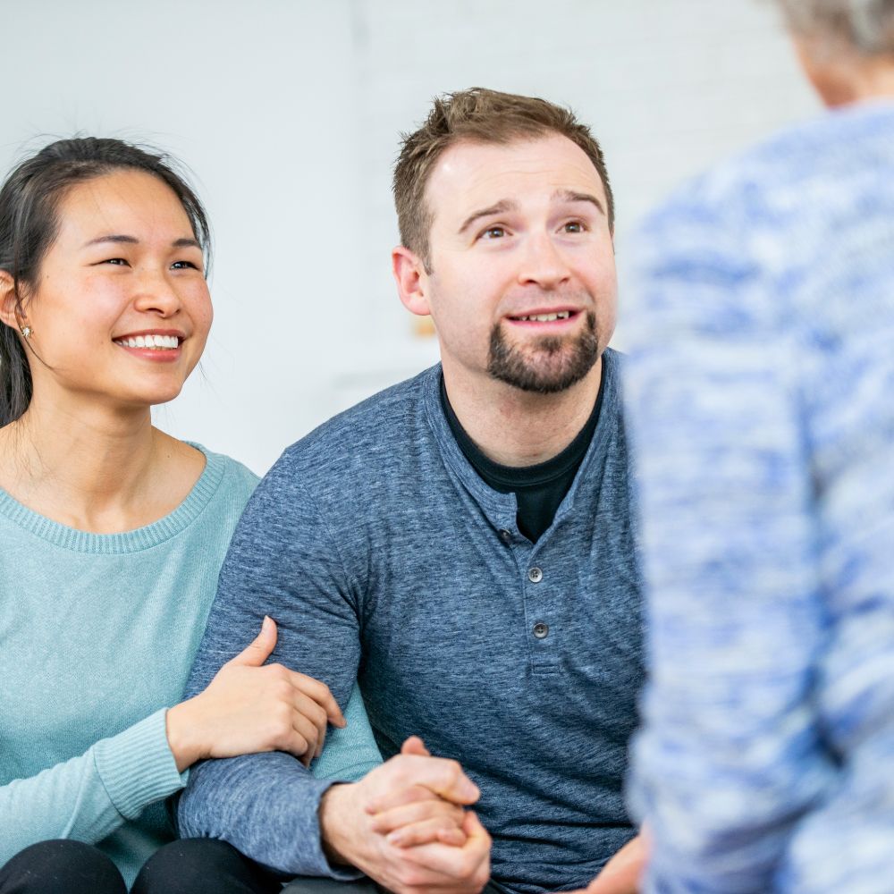 Smiling couple holding hands talking to counselor in bright room