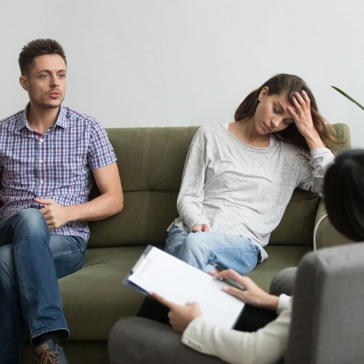 Couple sitting on a couch during counseling, with man speaking and woman holding her head in her hand