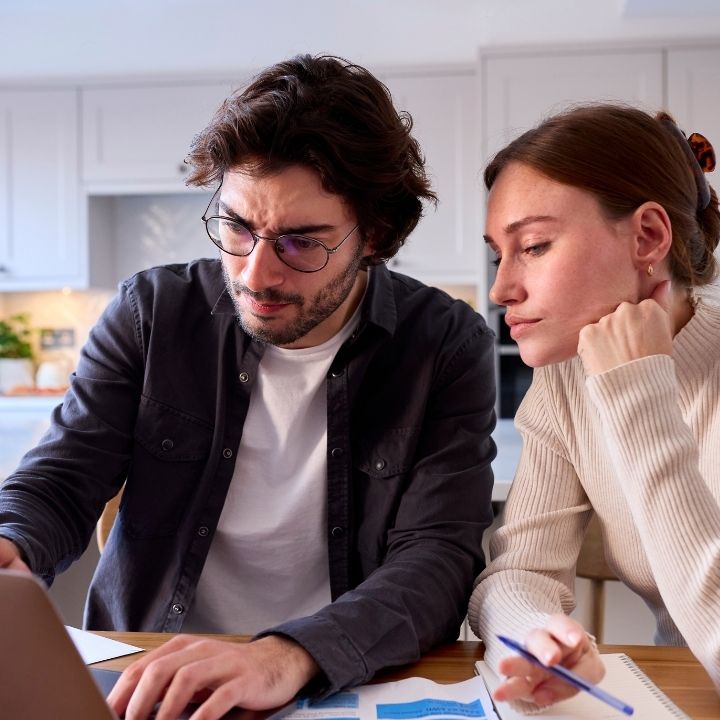 Couple working together at a kitchen table, focused on a laptop and paperwork while discussing something serious