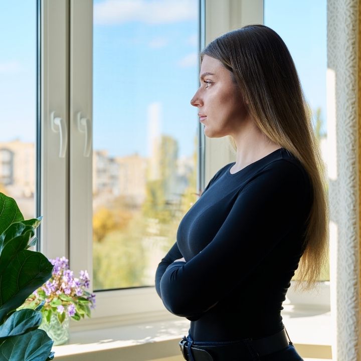 Woman standing by a window with her arms crossed, looking outside thoughtfully