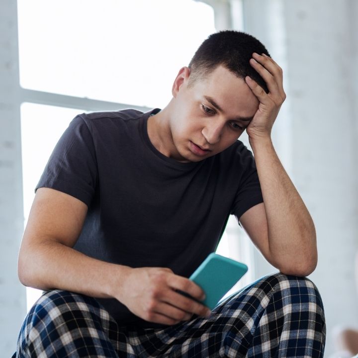 Man sitting on a chair, holding his head in distress while looking at his phone