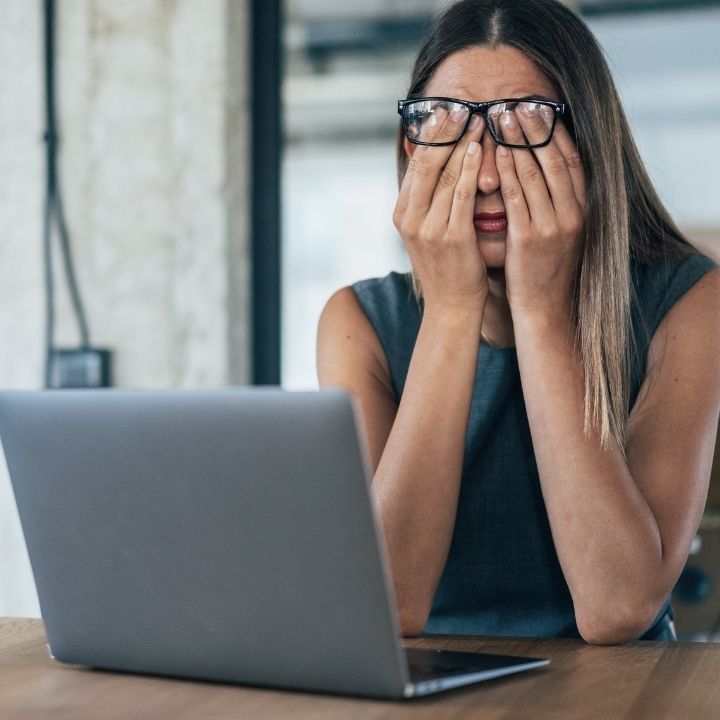 Woman sitting at a desk, covering her face with her hands in distress while looking at a laptop
