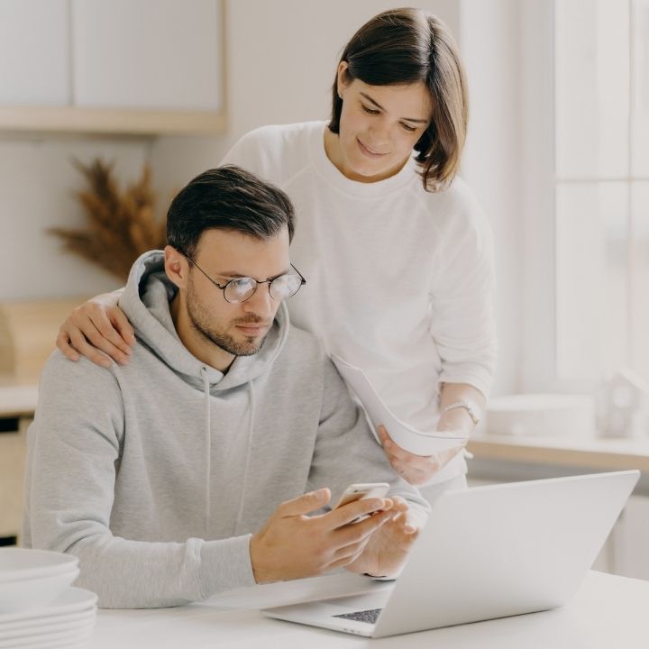 Woman standing behind a man at a kitchen table, both looking at a phone and laptop together