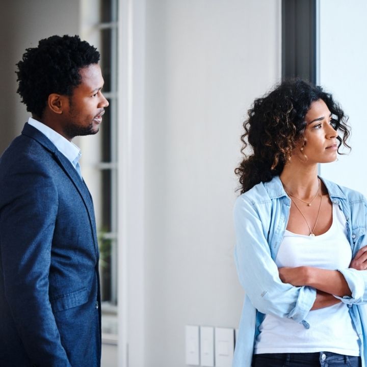 Couple standing apart indoors, one looking away with arms crossed while the other watches quietly.